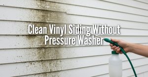 A close-up photograph of white vinyl siding being cleaned. The left side of the siding is covered in dark green and brown mildew, while the right side is clean. A hand is holding a green and white pump sprayer wand, directing a fine mist onto the dirty section to show the cleaning progress. Overlaid in the center is large, white, sans-serif text that reads: Clean Vinyl Siding Without Pressure Washer.