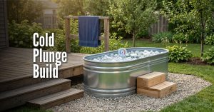 An image titled "Cold Plunge Build" showing a DIY outdoor cold plunge setup. It features a galvanized steel stock tank filled with water and floating ice cubes, sitting on a bed of light-colored gravel. A round thermometer is visible in the water, and small wooden steps are placed against the side for easy access. The setup is located next to a wooden deck with a blue towel hanging on a railing, surrounded by a green lawn and trees in a sunny backyard.