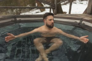 A bearded man sitting in an outdoor cold plunge tub or hot tub during winter with snow in the background.