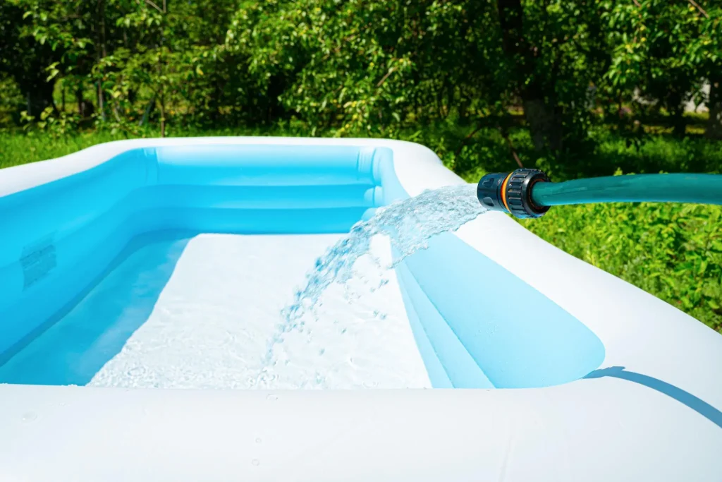 A green garden hose filling a blue and white inflatable pool with water in a sunny backyard with trees.