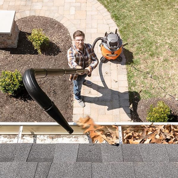 Homeowner using a wet dry vacuum with a curved extension tube to clean leaves out of gutters.