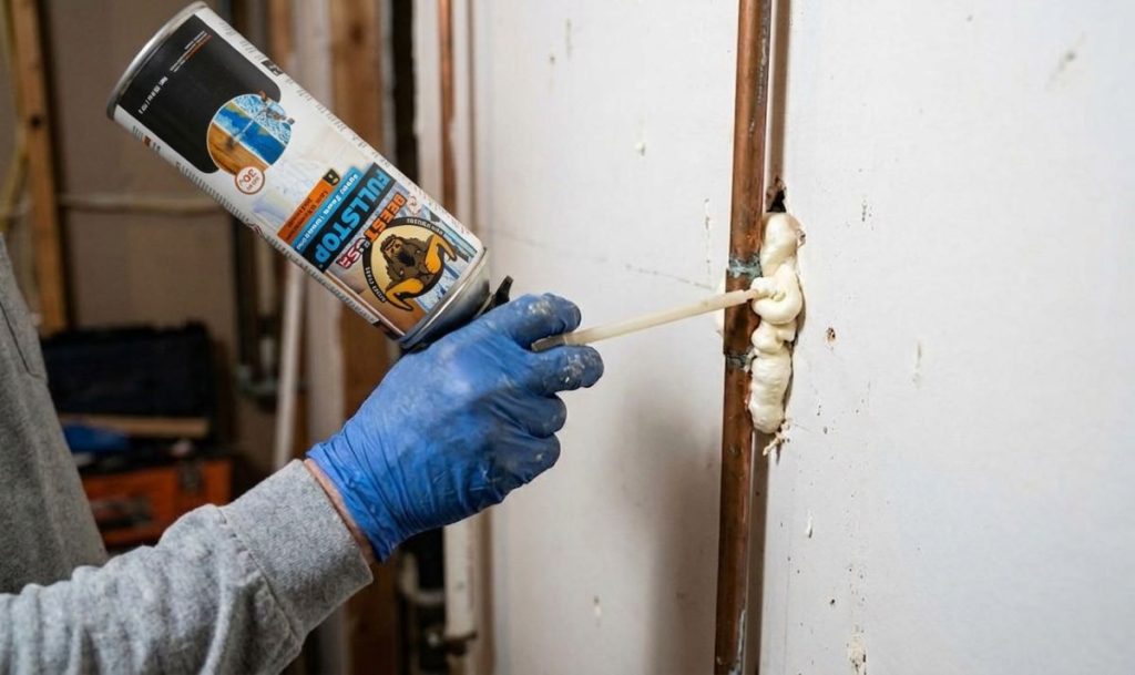 DIY homeowner using a spray foam insulation can to seal air leaks around copper pipes in an attic.
