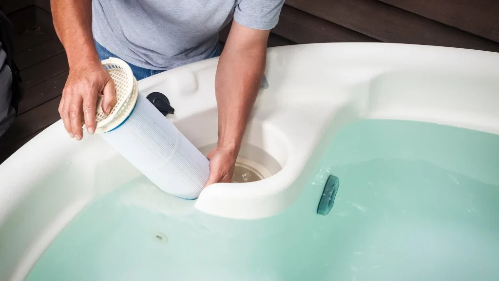 A close-up shot of a person’s hands removing or installing a large white cylindrical cartridge filter into a hot tub filled with clear blue water.