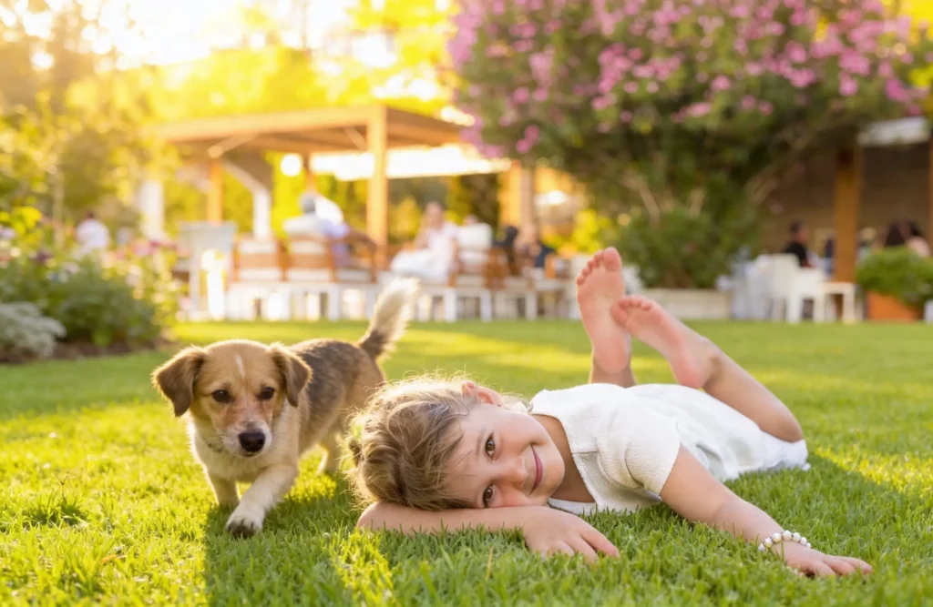 A girl in a white dress lying on green grass, smiling happily at the camera, with a small brown and white dog standing next to her in a sunlit garden.