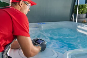 A technician in a red shirt and safety glasses using a handheld digital tablet to run diagnostics on a bubbling luxury hot tub.