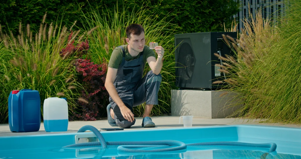 A pool maintenance technician in grey overalls kneeling by a blue swimming pool, carefully inspecting a water chemistry test kit. Large blue chemical containers and a pool heater are visible in the background.