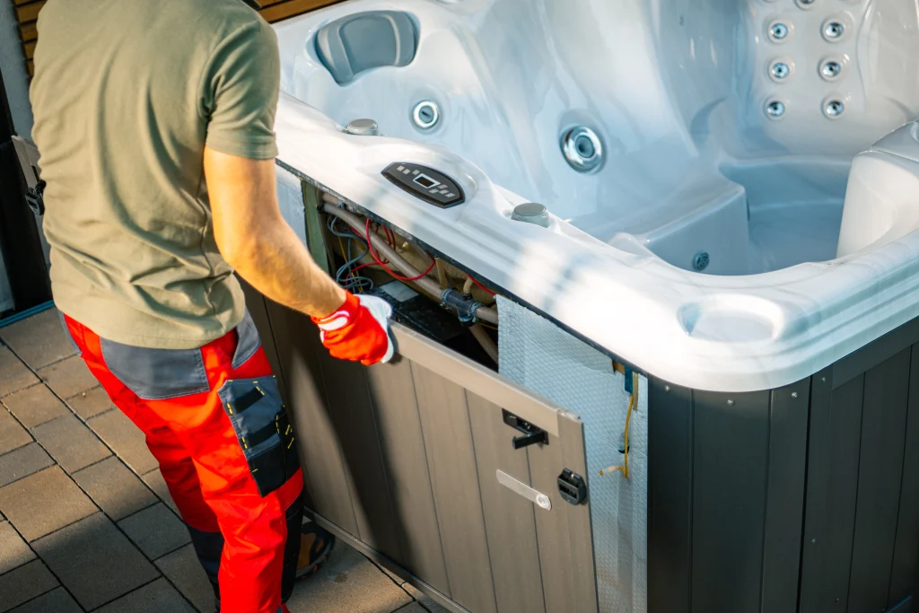 A technician in red work trousers and gloves removing the side panel of a modern white hot tub to access the internal wiring and plumbing for repair.